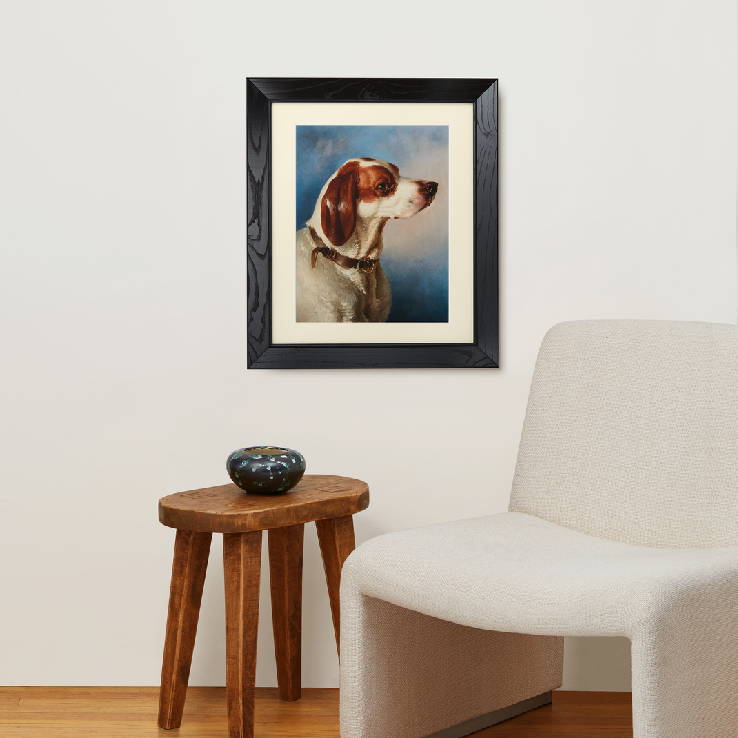 A dog portrait painting in a black ash angled frame hanging on a white wall next to a walnut table