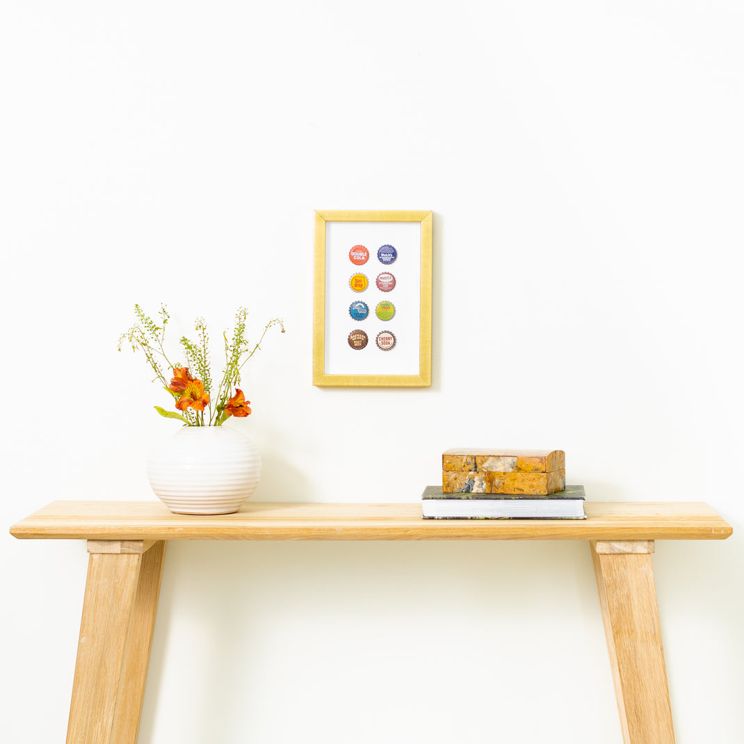 eight bottle caps framed in modern gold Dolly frame hanging on a white wall above a wood console table