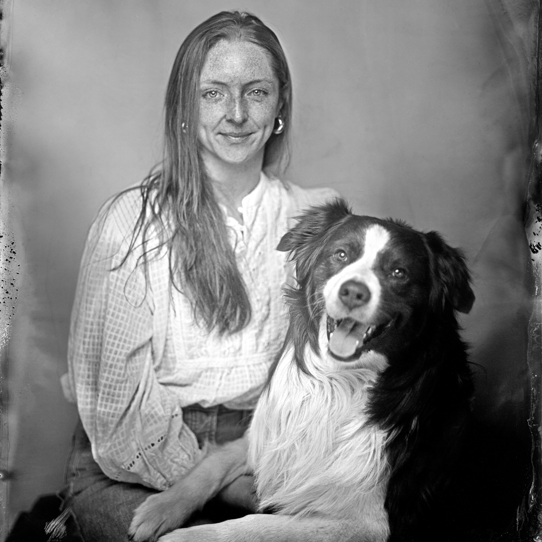 Black and white portrait of a woman seated beside a black-and-white dog against a mottled studio backdrop.