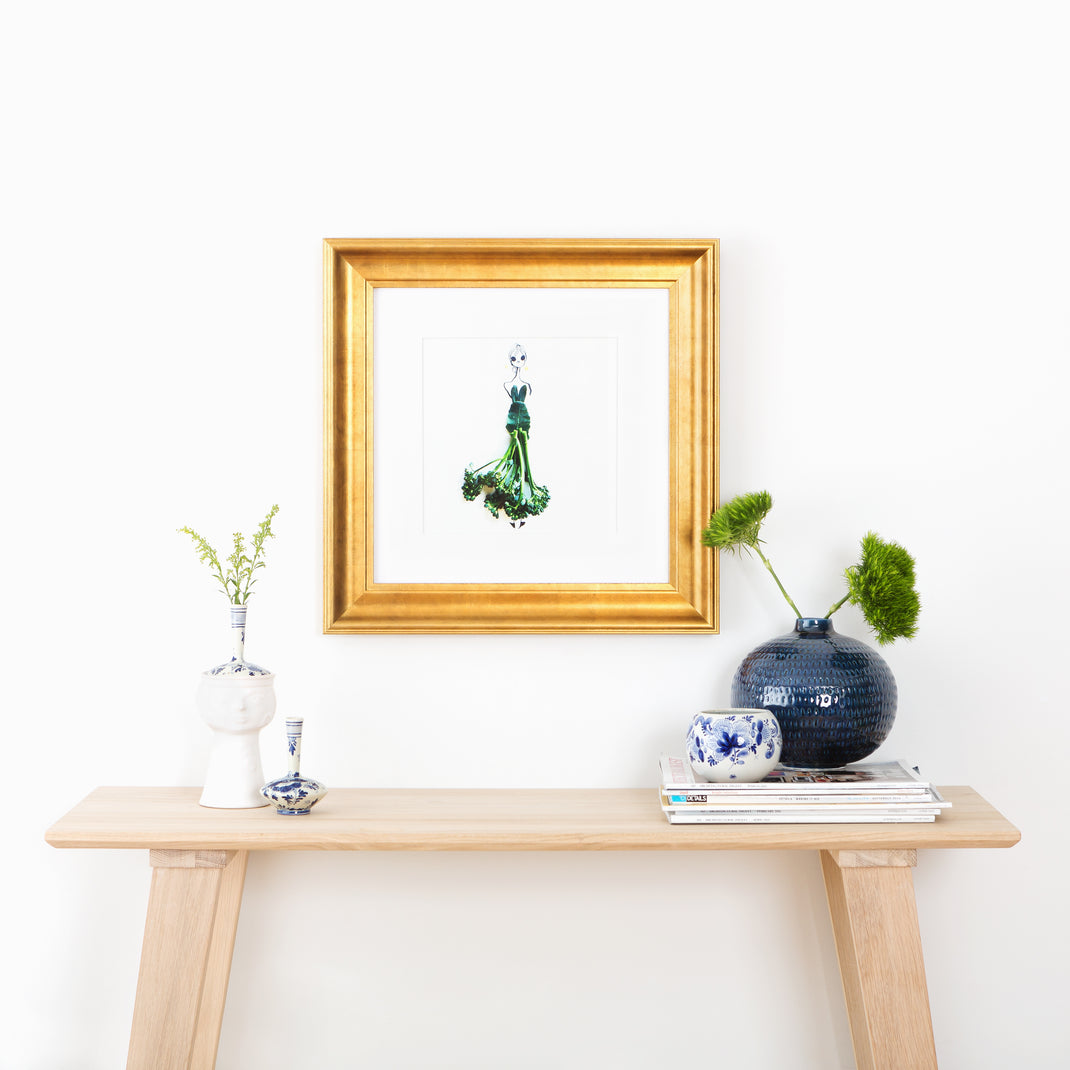 illustration of a woman with a broccolini skirt framed in wide sloped gold Chelsea frame with a white mat hanging on a white wall above a wooden console