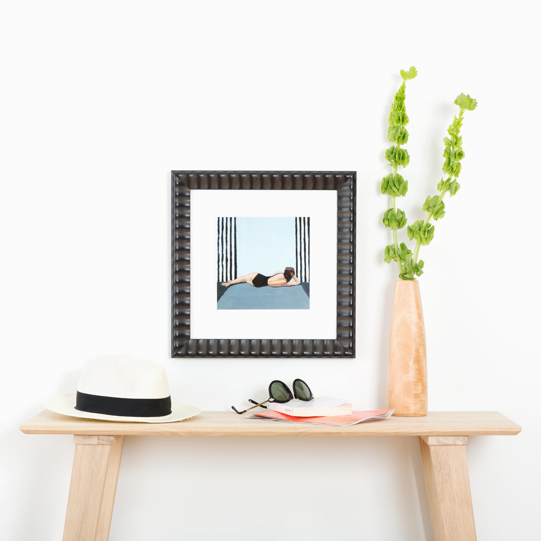abstract photo of a woman lounging in a black bathing suit framed in rippled Rialto frame with a white mat hanging on a white wall above a wood console table
